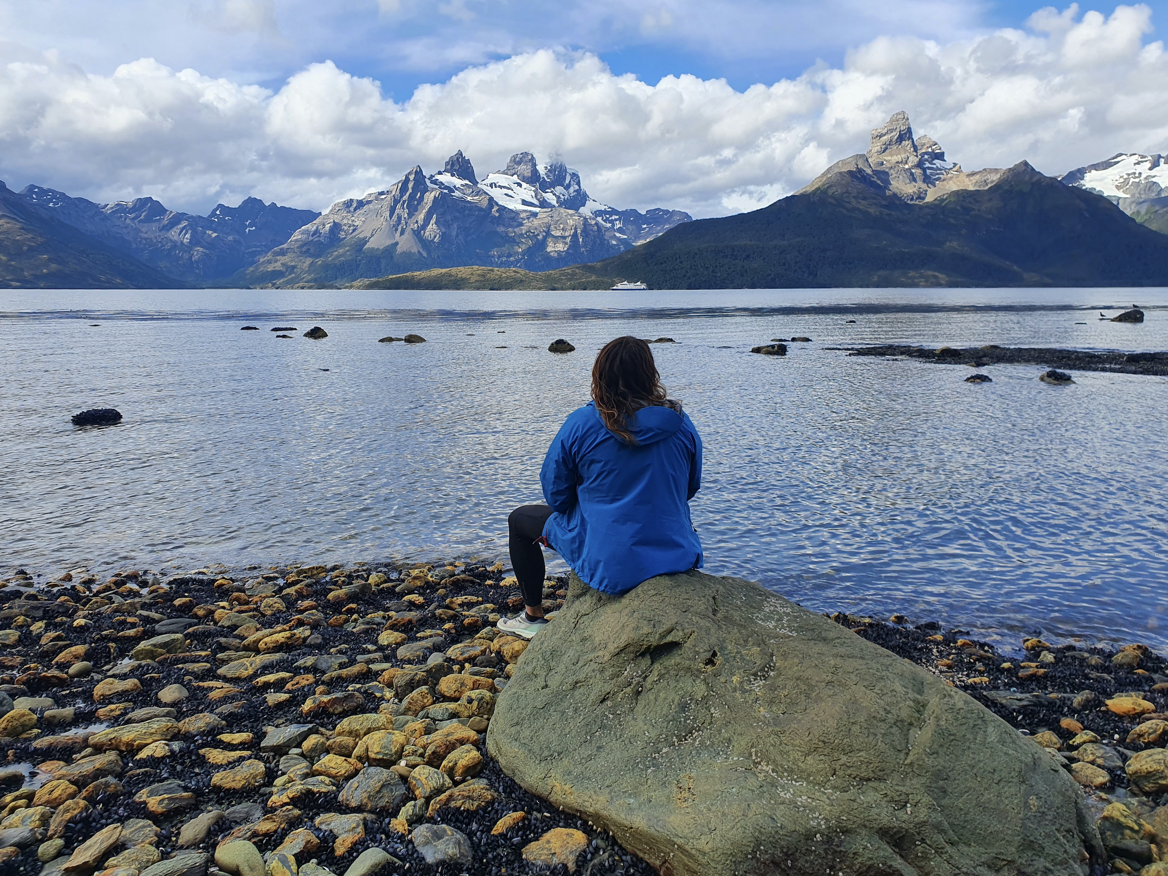 A tourist sits on a rock facing the sea and distant mountains of Tierra del Fuego