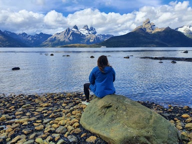 A tourist sits on a rock facing the sea and distant mountains of Tierra del Fuego